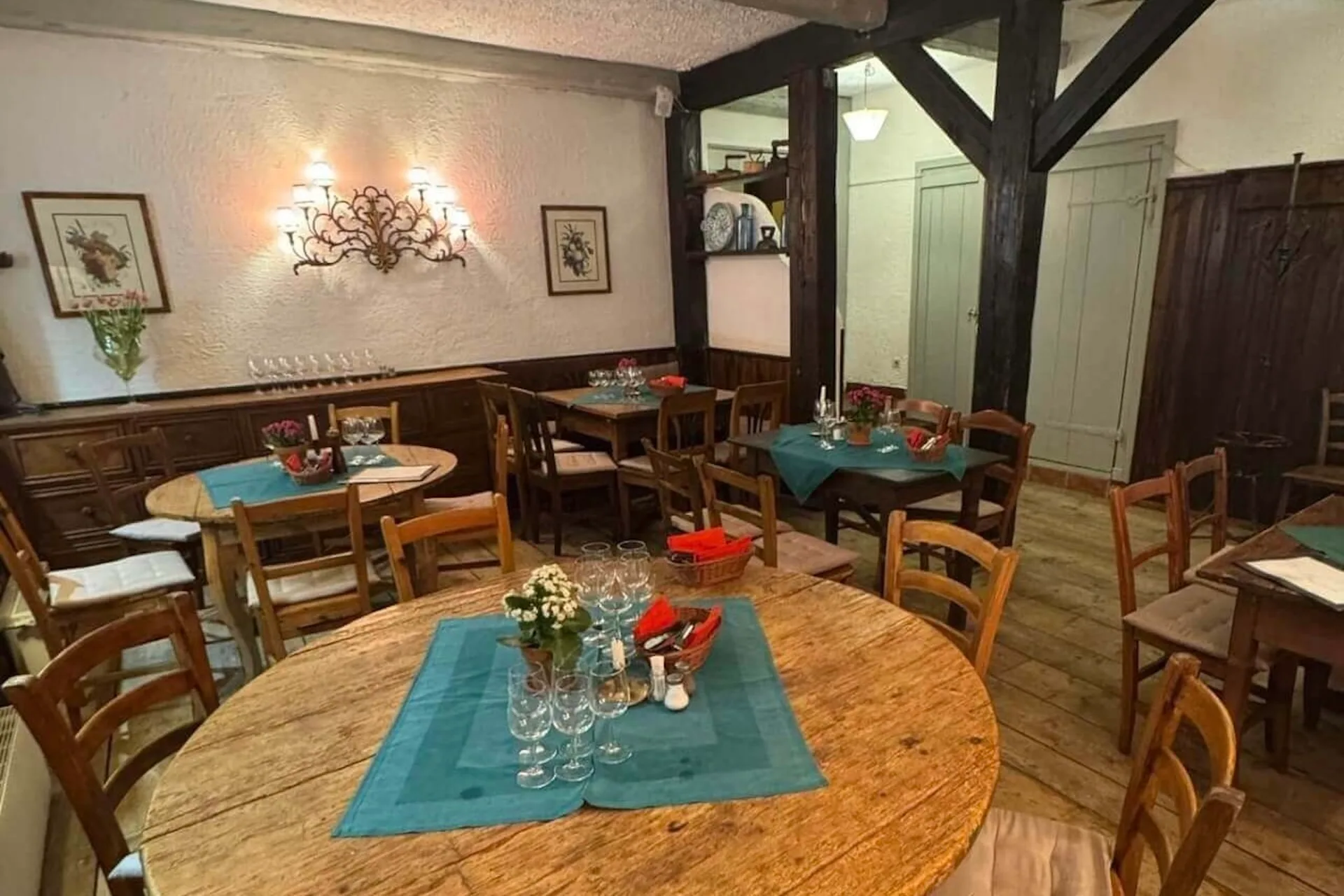 Ein gemütlicher Restaurantbereich mit Holztischen und -stühlen, türkisfarbenen Tischtüchern, ordentlich arrangierten Gläsern und Blumensträußen. Die warme Beleuchtung und die rustikale Einrichtung schaffen eine einladende Atmosphäre.