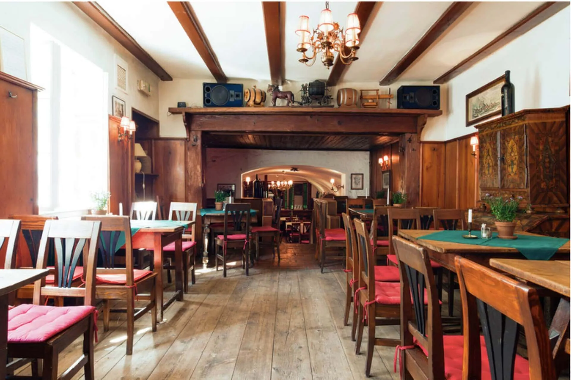 Ein gemütliches Restaurant mit Holzfußböden, Tischen und Stühlen mit roten Polstern. Pflanzen schmücken die Tische, und durch ein Fenster auf der linken Seite strömt Sonnenlicht. Ein Kronleuchter hängt von einer Balkendecke.