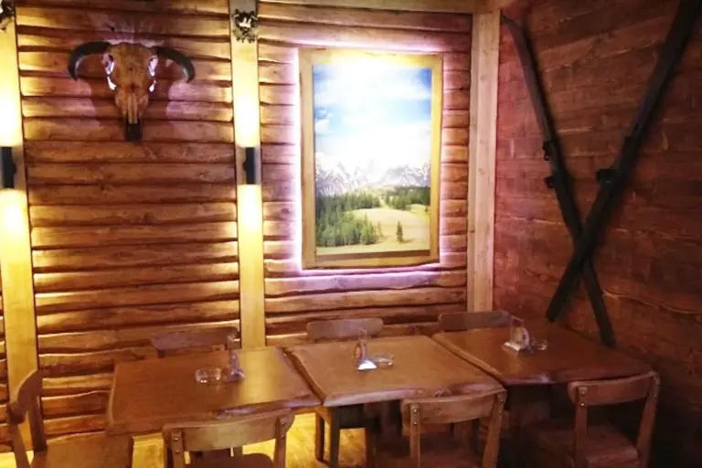 Ein rustikaler hölzerner Essbereich mit drei Tischen, Holzstühlen, einem Gemälde einer Berglandschaft an der Wand, einem Stierschädel und zwei gekreuzten schwarzen Objekten an den Holzwänden.