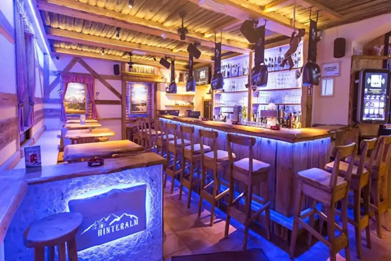 Eine gemütliche, rustikale Bar mit Holzbalken, hohen Hockern und einer blau beleuchteten Theke mit der Aufschrift Hinteralm. Flaschenregale säumen die Rückwand, und die weiche, warme Beleuchtung verleiht dem Raum ein einladendes Hüttengefühl.