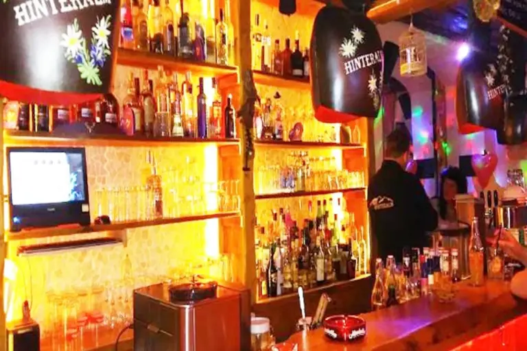Eine hell erleuchtete Bar mit Regalen voller Spirituosenflaschen und Glaswaren, die von warmem gelbem und orangefarbenem Licht angestrahlt werden. Hinter dem Tresen steht ein Barkeeper, im Vordergrund stehen Barhocker und Bierzapfanlagen.