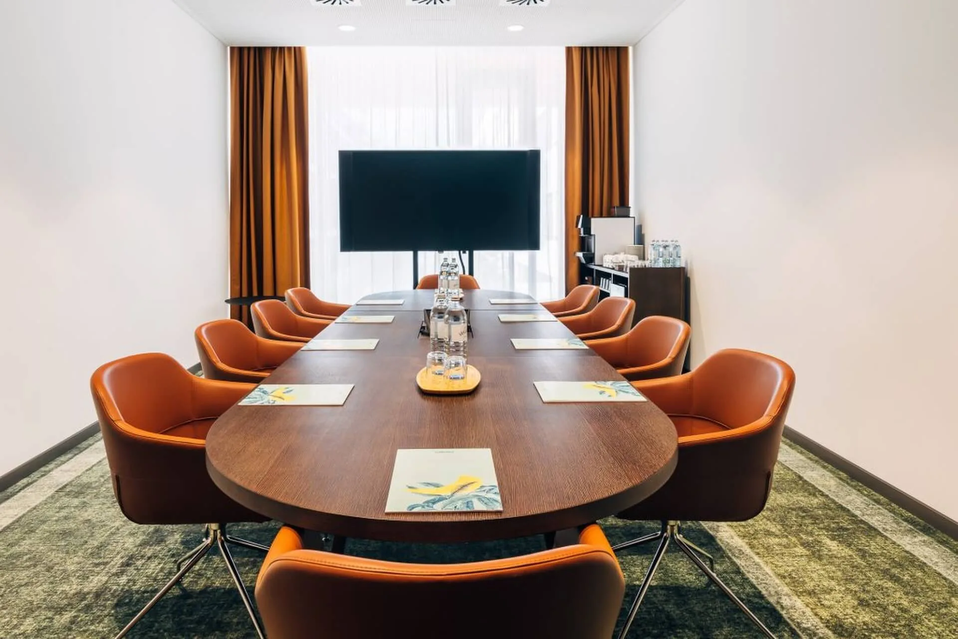 Ein moderner Konferenzraum mit einem langen Holztisch, umgeben von orangefarbenen Stühlen, die mit Notizblöcken und Wasserflaschen ausgestattet sind, vor einem großen Fernsehbildschirm. Gelbe Vorhänge umrahmen ein helles Fenster am Ende des Raums.