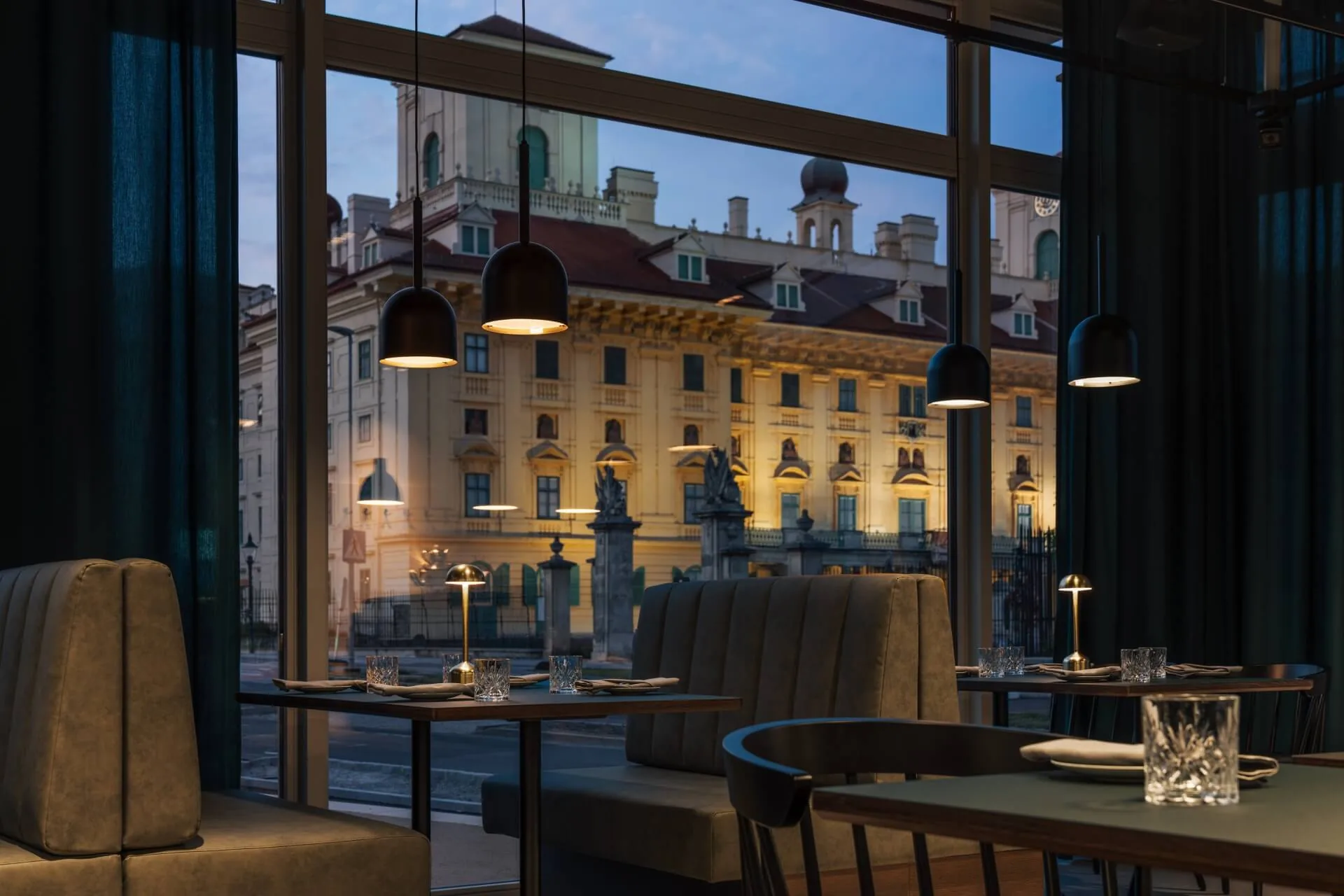 A dimly lit restaurant with modern decor and upholstered booths overlooks a grand, illuminated historic building through large windows at dusk.