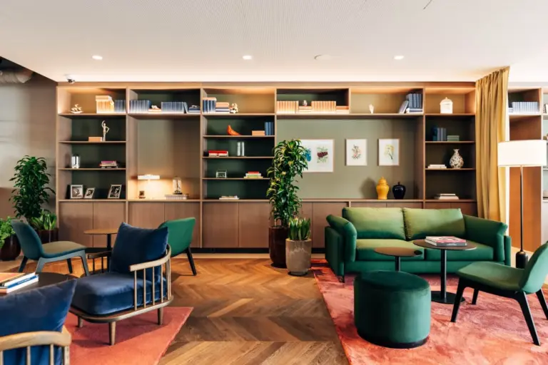 Modernes Wohnzimmer mit grünem Sofa, Sesseln, runder Ottomane und mehreren Topfpflanzen. Bücherregale mit Büchern, Dekorationsobjekten und gerahmter Kunst säumen die Rückwand. Holzboden und bunte Teppiche vervollständigen den gemütlichen Raum.