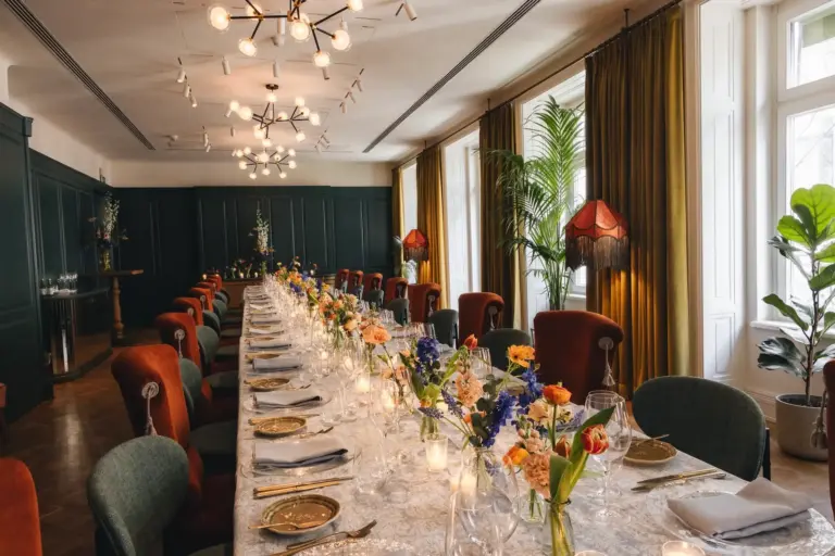 Bel Étage Hotel Motto In einem stilvollen Raum mit Samtstühlen, grünen Wänden, großen Fenstern und Vintage-Lampen ist ein langer, eleganter Esstisch mit Blumen, Kerzen und Gläsern gedeckt.