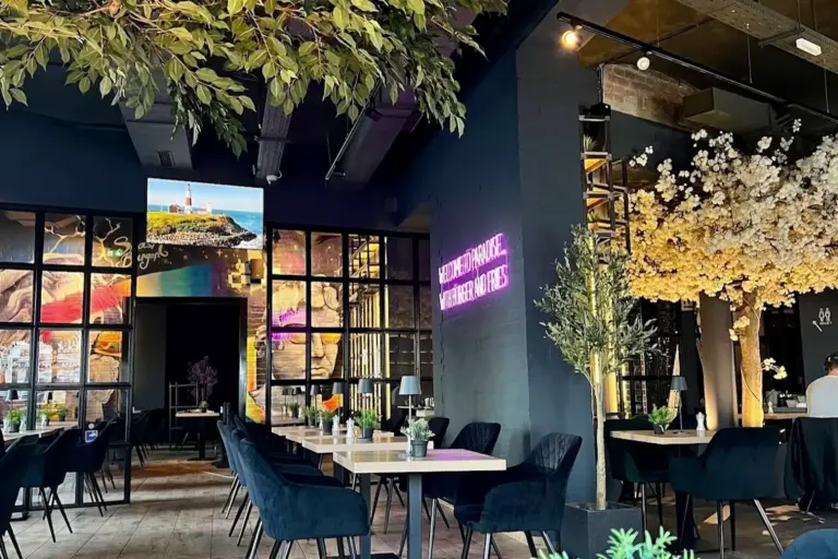 Modernes Restaurant mit dunkelblauen Wänden, Leuchtreklame, Samtstühlen, Holztischen, großen Fenstern und Zimmerpflanzen, darunter ein Baum mit weißen Blumen und von der Decke hängendem Grünzeug. Warme, einladende Atmosphäre.