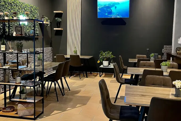 Modernes Cafe mit Holztischen und braunen Stühlen, dekoriert mit Topfpflanzen und Regalen. Ein Fernseher an einer dunklen Wand zeigt ein landschaftliches Bild. Die Atmosphäre ist gemütlich und einladend mit einer Mischung aus natürlichem und modernem Dekor.