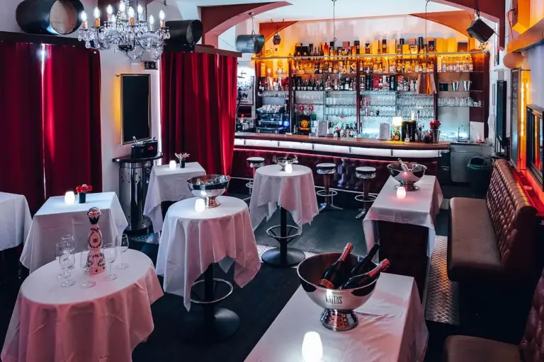 Eine stilvolle Lounge-Bar mit weiß gedeckten Tischen, Champagnerflaschen in Eiskübeln, leuchtenden Tischlampen, roten Vorhängen, einem Kronleuchter und einer gut gefüllten Bar im Hintergrund.