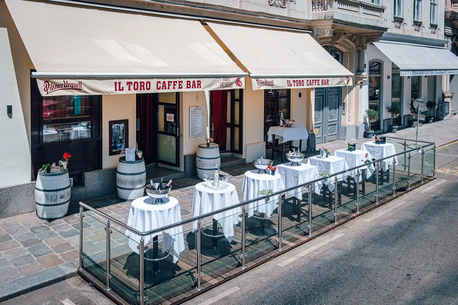 Außenbereich der IL TORO CAFFE BAR mit runden Tischen mit weißen Tischtüchern und Metallstühlen auf einer Holzterrasse entlang einer Stadtstraße, mit Weinfässern als Dekoration und einem beigen Vordach.
