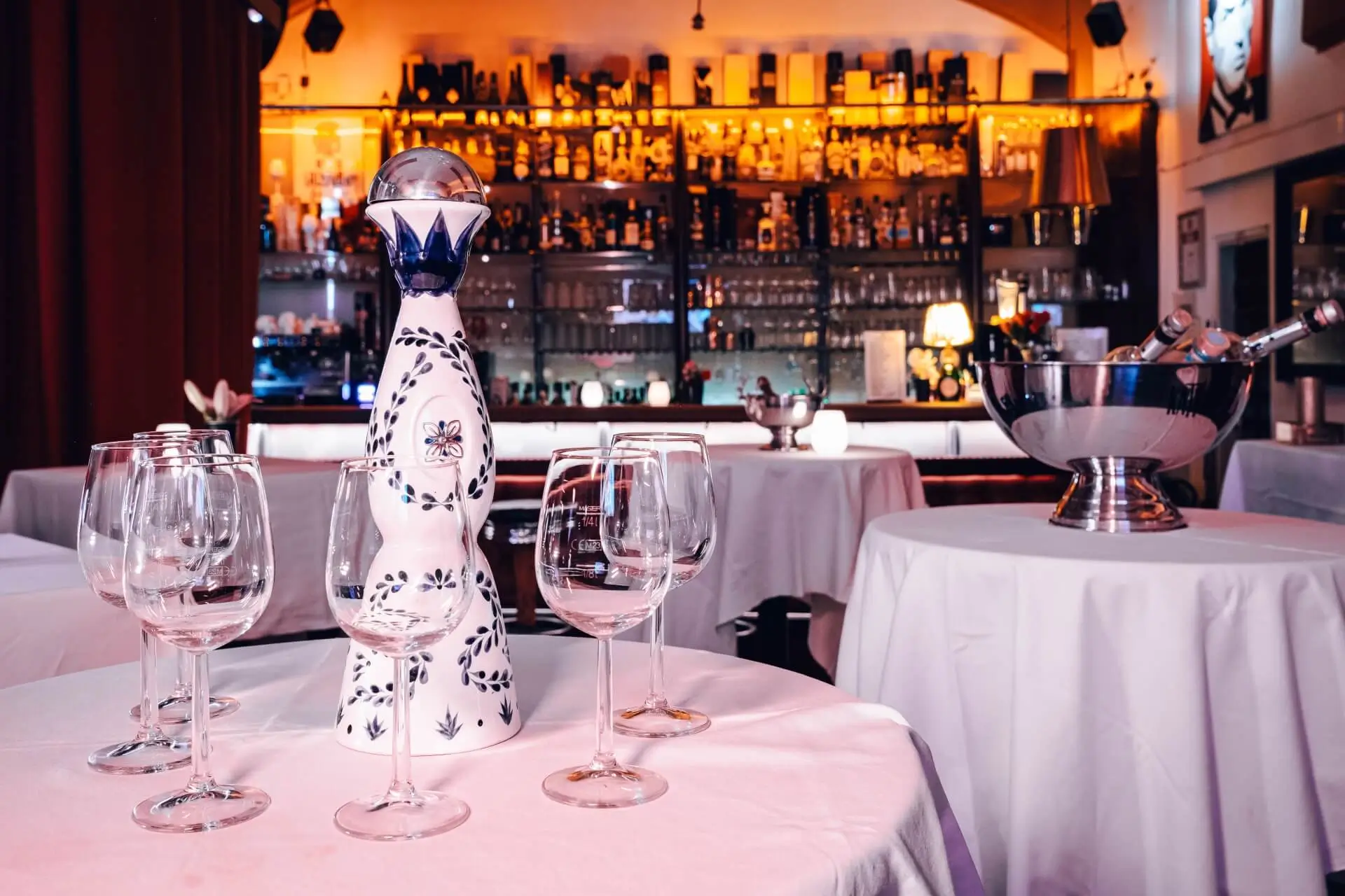 Ein Tisch mit leeren Weingläsern und einer dekorativen Tequilaflasche steht in einer stilvollen Bar mit weißen Tischtüchern, einem großen beleuchteten Spirituosenregal und einem Champagnerkübel im Hintergrund.