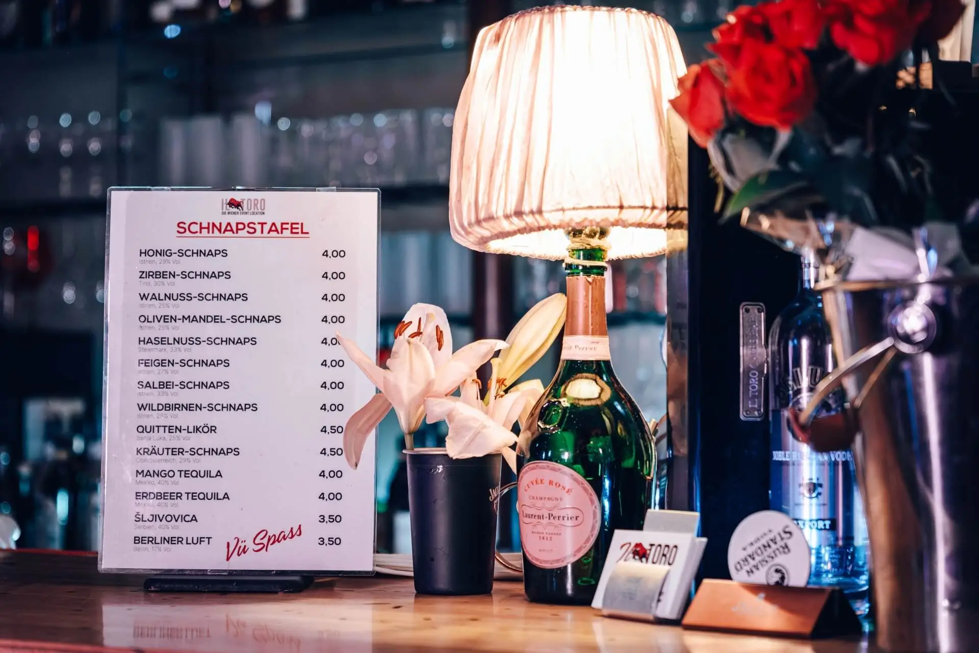 Eine Bartheke mit einer Getränkekarte, einer Blume in einem schwarzen Topf, einer Flasche Champagner, einer beleuchteten Tischlampe und einem Strauß roter Rosen vor einem unscharfen Hintergrund aus Glaswaren.