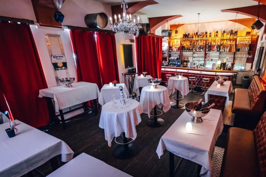 Ein gemütliches Restaurant mit weiß gedeckten Tischen, roten Vorhängen, einem Kronleuchter und einer gut gefüllten Bar im Hintergrund. Die Beleuchtung ist warm und einladend.