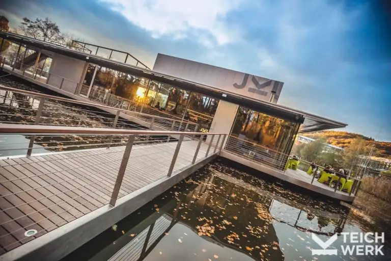JKU Teichwerk Modernes Gebäude auf einer Plattform im See mit einer Sitzecke im Freien, das über eine Holzbrücke mit dem Ufer verbunden ist.