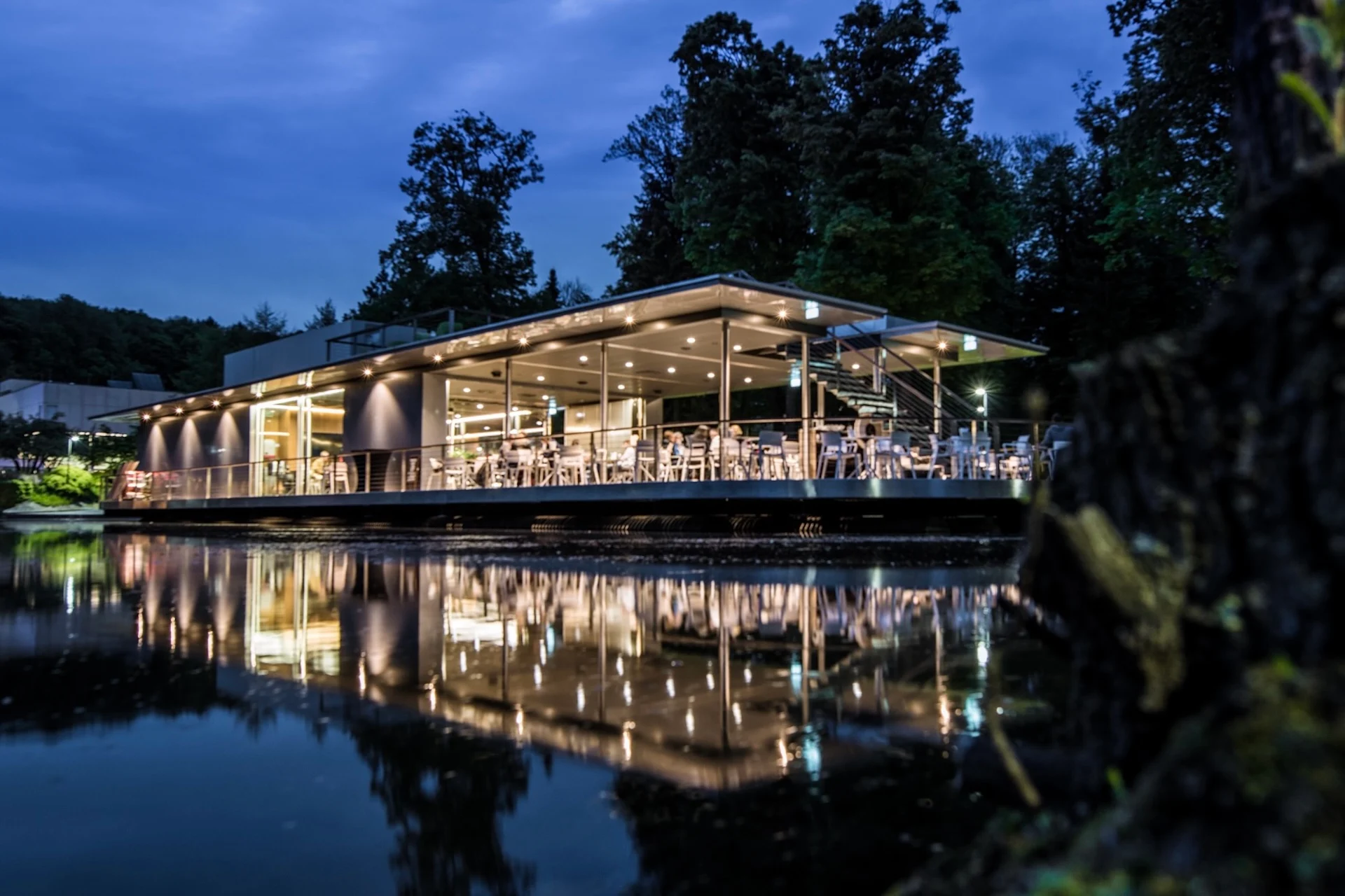 Ein modernes Restaurant mit Glaswänden an einem See in der Abenddämmerung, dessen Innenbeleuchtung sich im Wasser spiegelt.