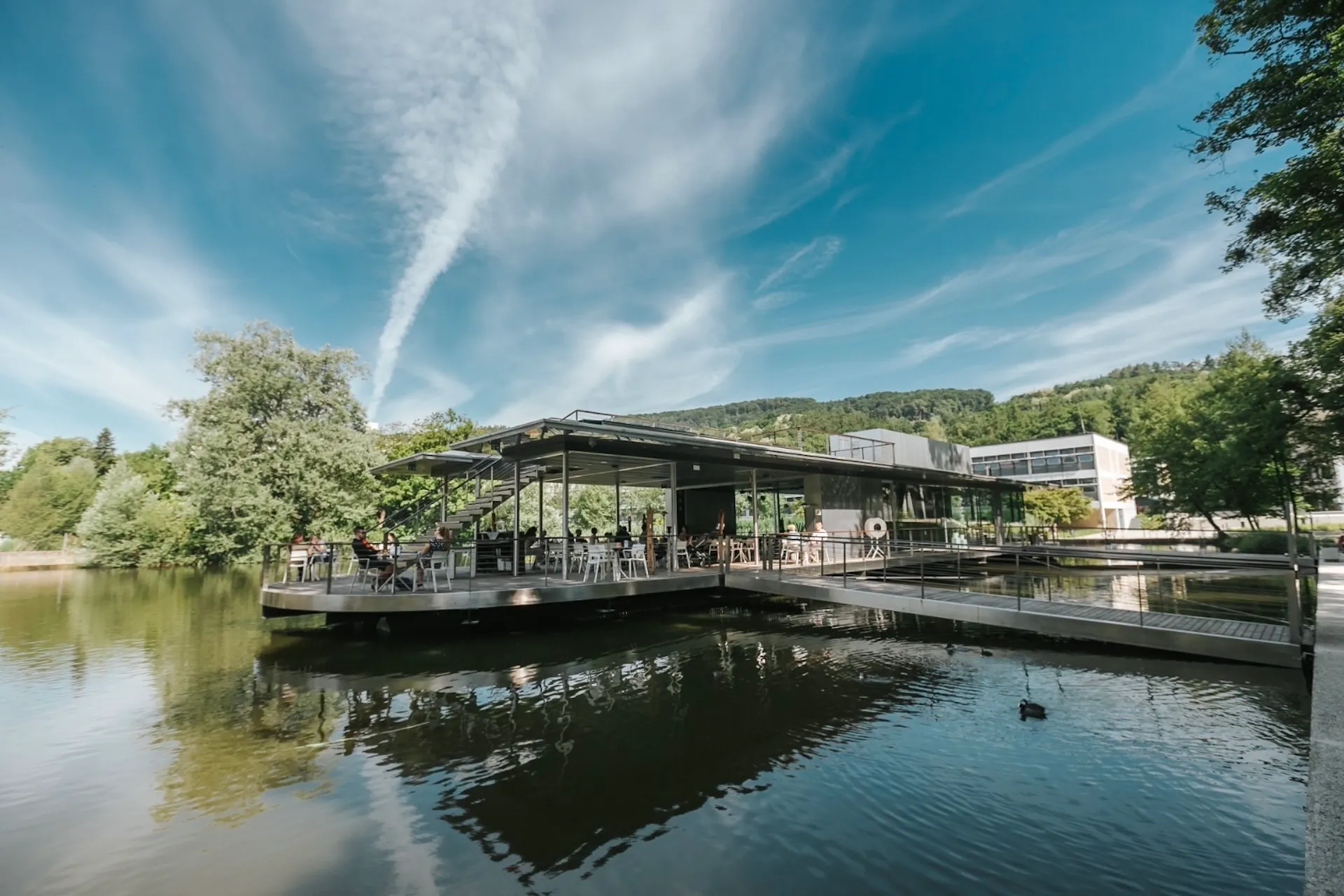 Ein modernes Restaurantgebäude mit Flachdach auf einer Plattform über einem See, das über einen Steg mit dem Ufer verbunden ist, auf dessen Terrasse Gäste speisen.