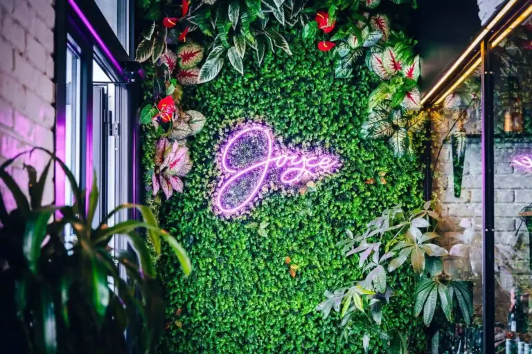 Eine rosafarbene Leuchtreklame mit der Aufschrift Joyce ist an einer üppigen grünen Wand angebracht, die mit verschiedenen Pflanzen und kleinen roten Blumen bedeckt ist und eine lebendige, tropische Atmosphäre schafft.