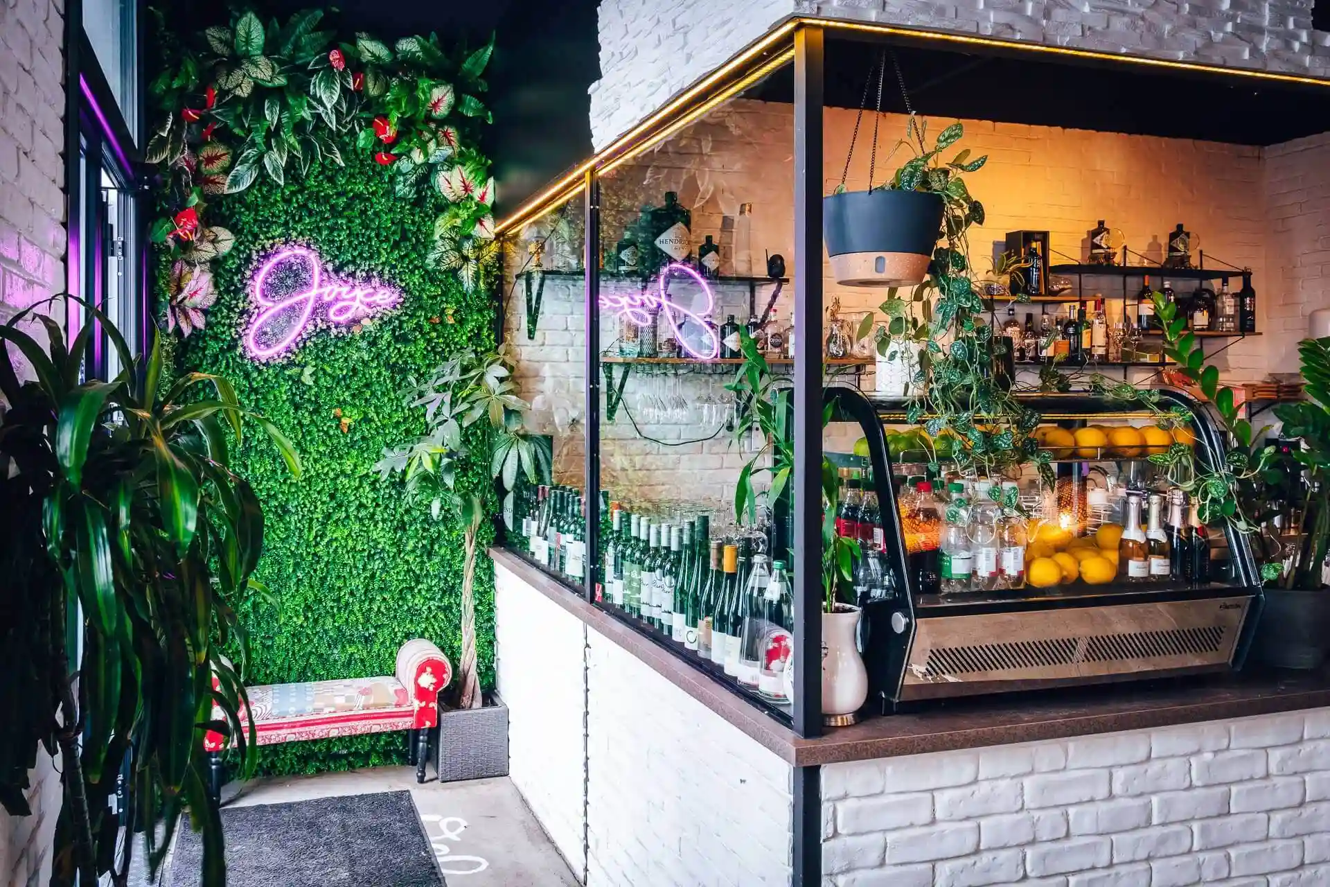Ein modernes Café mit einer weißen Backsteintheke, hängenden Pflanzen, Regalen mit Flaschen und Gläsern und einer üppig grünen Wand mit einer rosa Leuchtreklame. Eine kleine rote Bank steht neben dem Eingang.