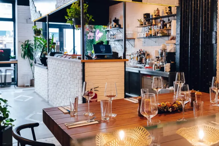 Modernes Restaurantinterieur mit einem Holztisch, der mit Gläsern, Besteck und einem Tafelaufsatz gedeckt ist, vor einem Barbereich mit Regalen mit Flaschen und Pflanzen, und Sonnenlicht, das durch große Fenster einfällt.