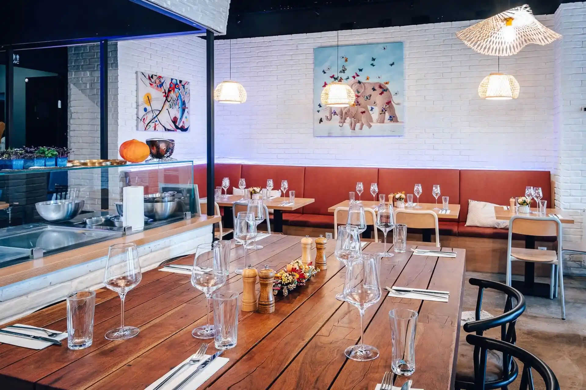 Ein modernes Restaurant mit Holztischen, die mit Gläsern und Geschirr gedeckt sind, orangefarbenen Sitzkissen, bunten Kunstwerken an weißen Backsteinwänden und Hängelampen, die eine gemütliche Atmosphäre schaffen.