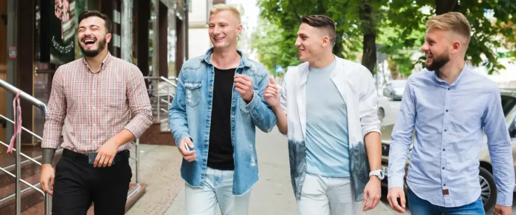 Vier junge Männer gehen an einem sonnigen Tag in Wien spazieren und lachen zusammen. Sie scheinen einen lebhaften Junggesellenabschied zu genießen, während sie an Bäumen und Gebäuden vorbeischlendern.