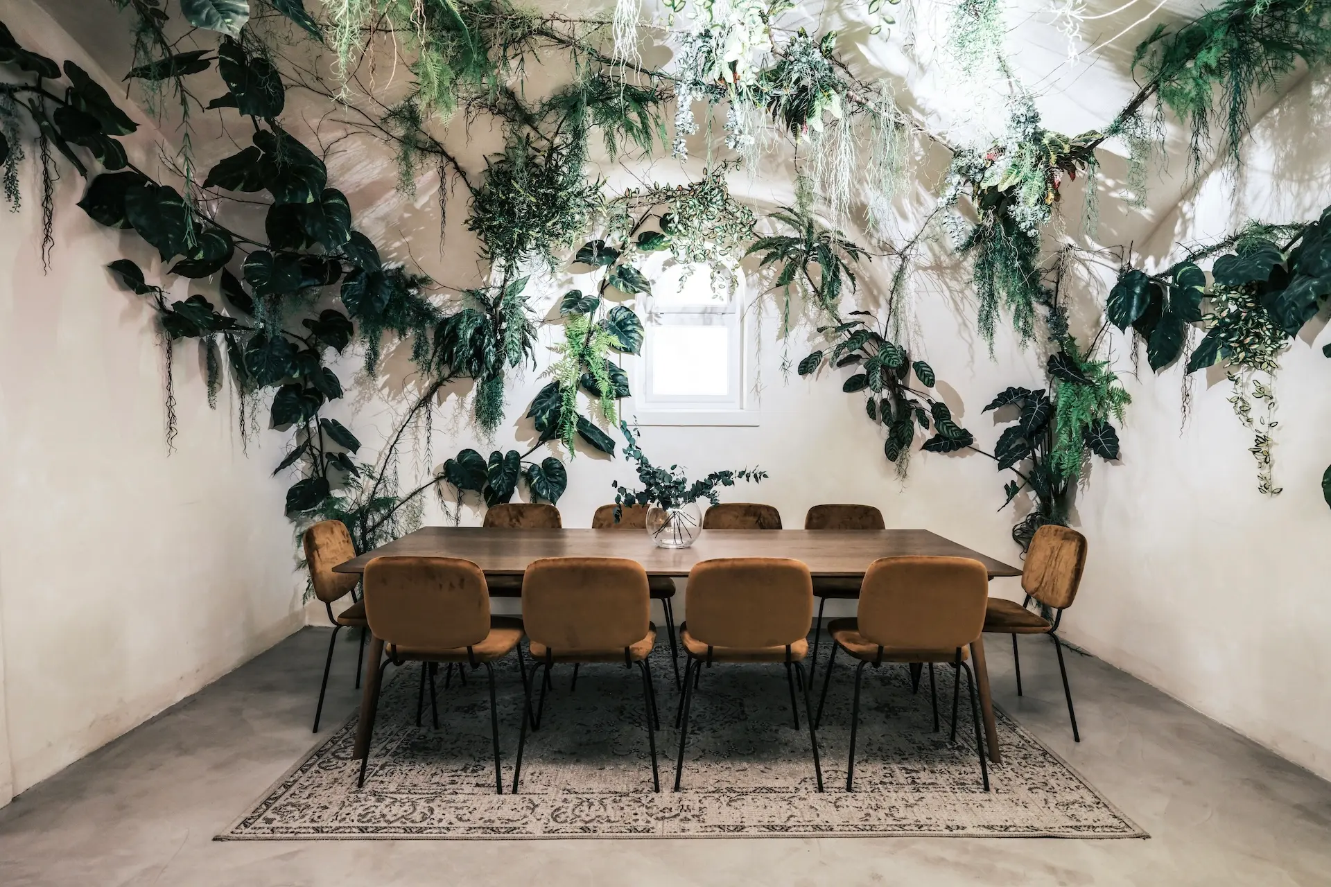 KLYO Graz Ein Esszimmer mit einem langen Holztisch, acht braunen Stühlen, einem gemusterten Teppich und einer weißen Wand, die mit üppigen grünen Pflanzen bedeckt ist, mit einem kleinen Fenster in der Mitte.