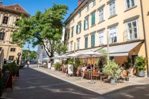 Eine sonnige Straßenszene mit Menschen, die an Tischen im Freien unter Sonnenschirmen vor einem gelben Gebäude mit grünen Fensterläden speisen. Üppige Bäume und Topfpflanzen säumen den kopfsteingepflasterten Gehweg.