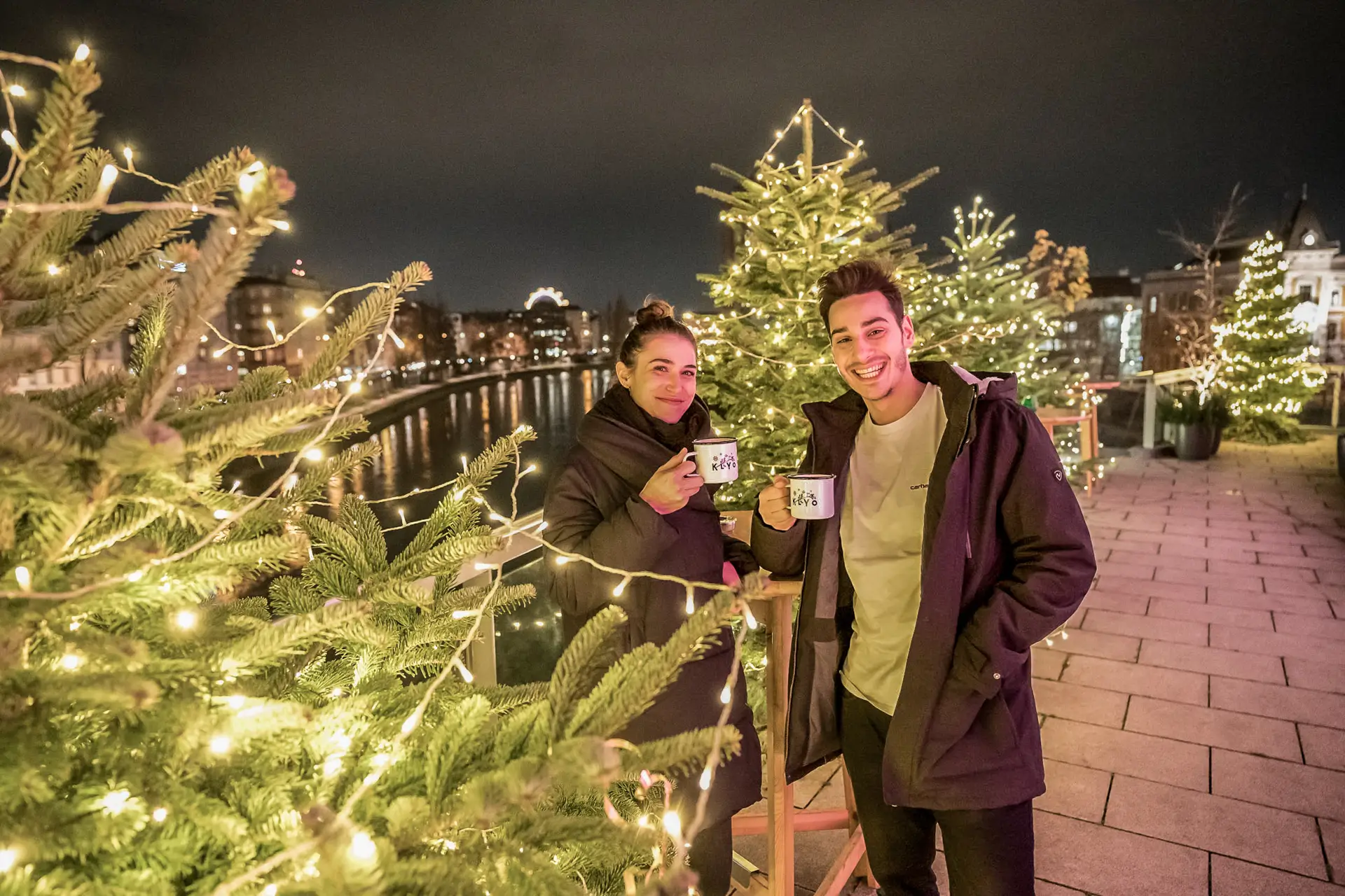 Eine Frau und ein Mann stehen nachts im Freien, lächeln und halten Tassen inmitten von geschmückten Weihnachtsbäumen mit Lichterketten an einem Fluss, der von städtischen Gebäuden gesäumt ist.