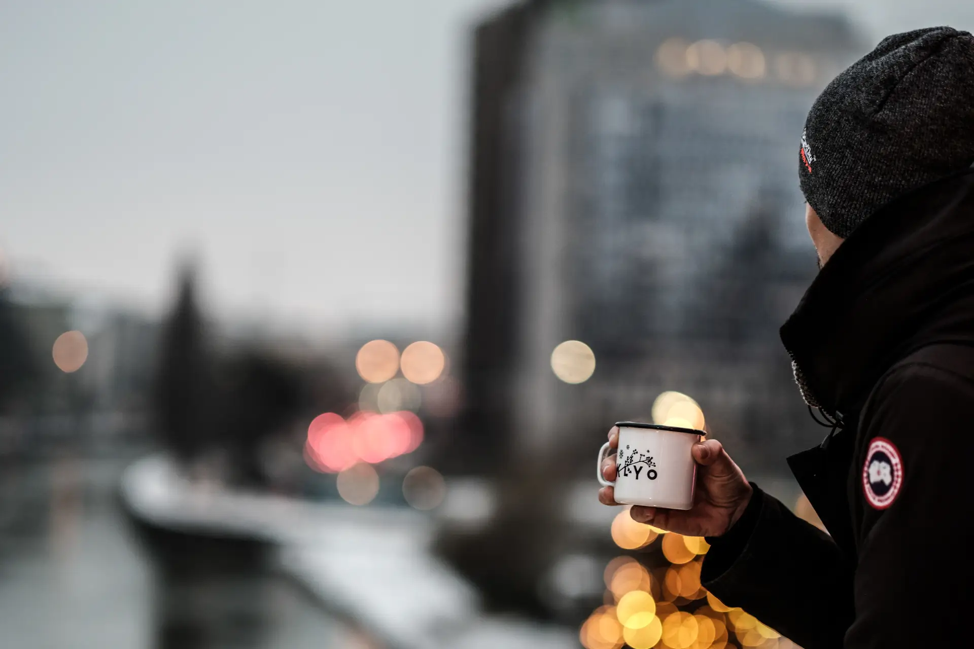 Eine Person in dunkler Jacke und Mütze hält einen weißen Becher mit schwarzem Text in der Hand. Sie steht im Freien in der Nähe eines verschwommenen Stadthintergrunds mit Bokeh-Lichtern und einem Fluss.