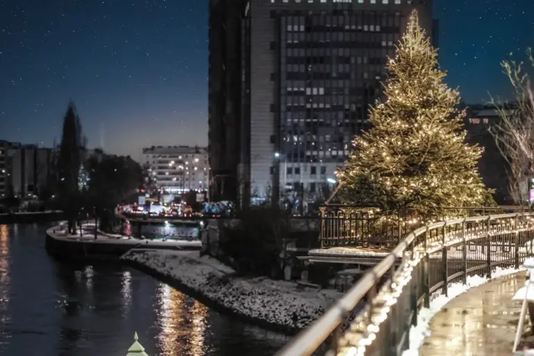 Ein geschmückter Weihnachtsbaum mit hellen Lichtern steht an einem nächtlichen Fluss, in dem sich die Gebäude und Straßenlaternen der Stadt auf dem Wasser spiegeln und Schnee den Boden bedeckt.