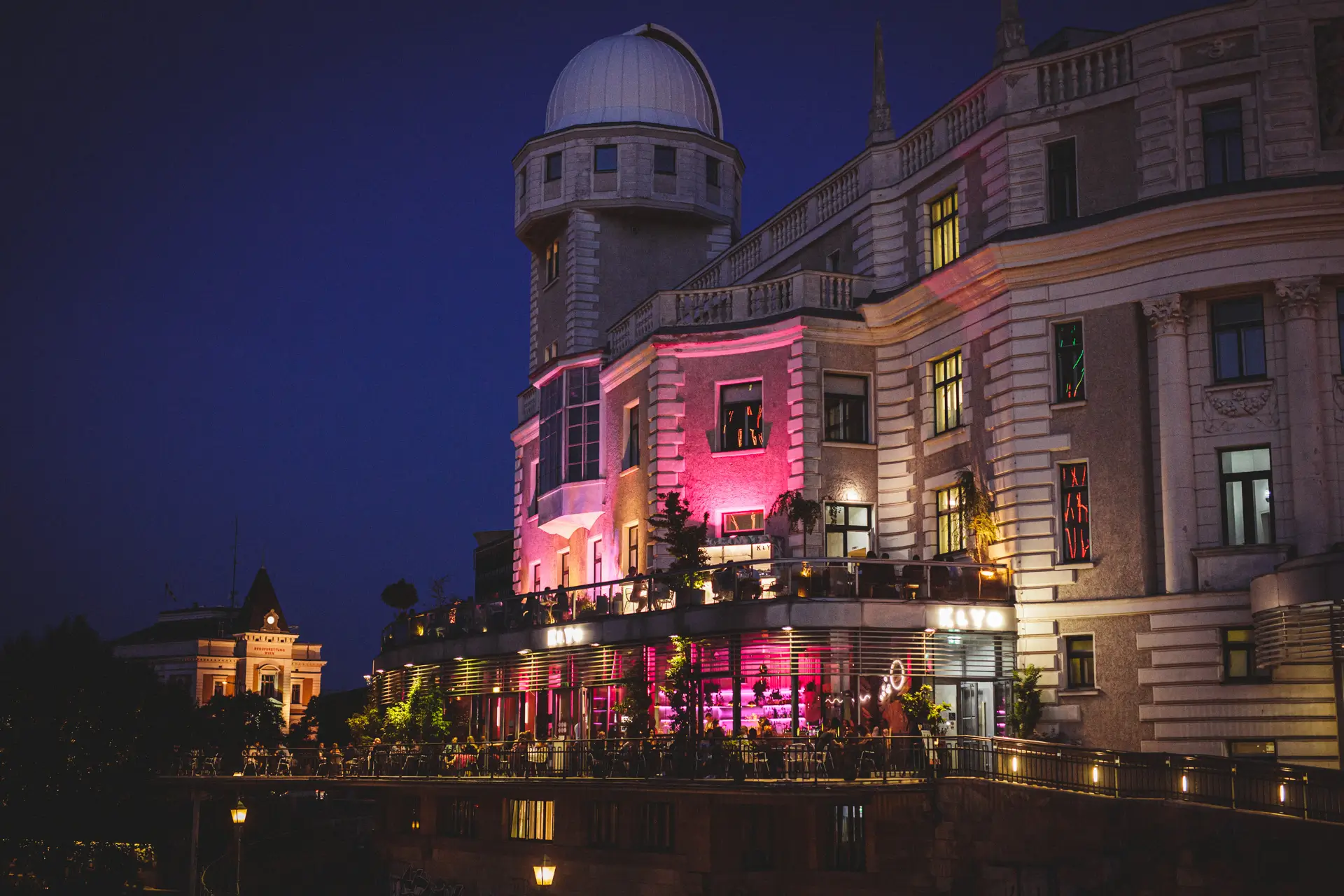 Ein großes historisches Gebäude mit einer Kuppel und einem Observatorium, das nachts mit rosafarbenen und violetten Lichtern beleuchtet wird und auf dessen Terrasse man mit Blick auf die Stadt speisen kann.
