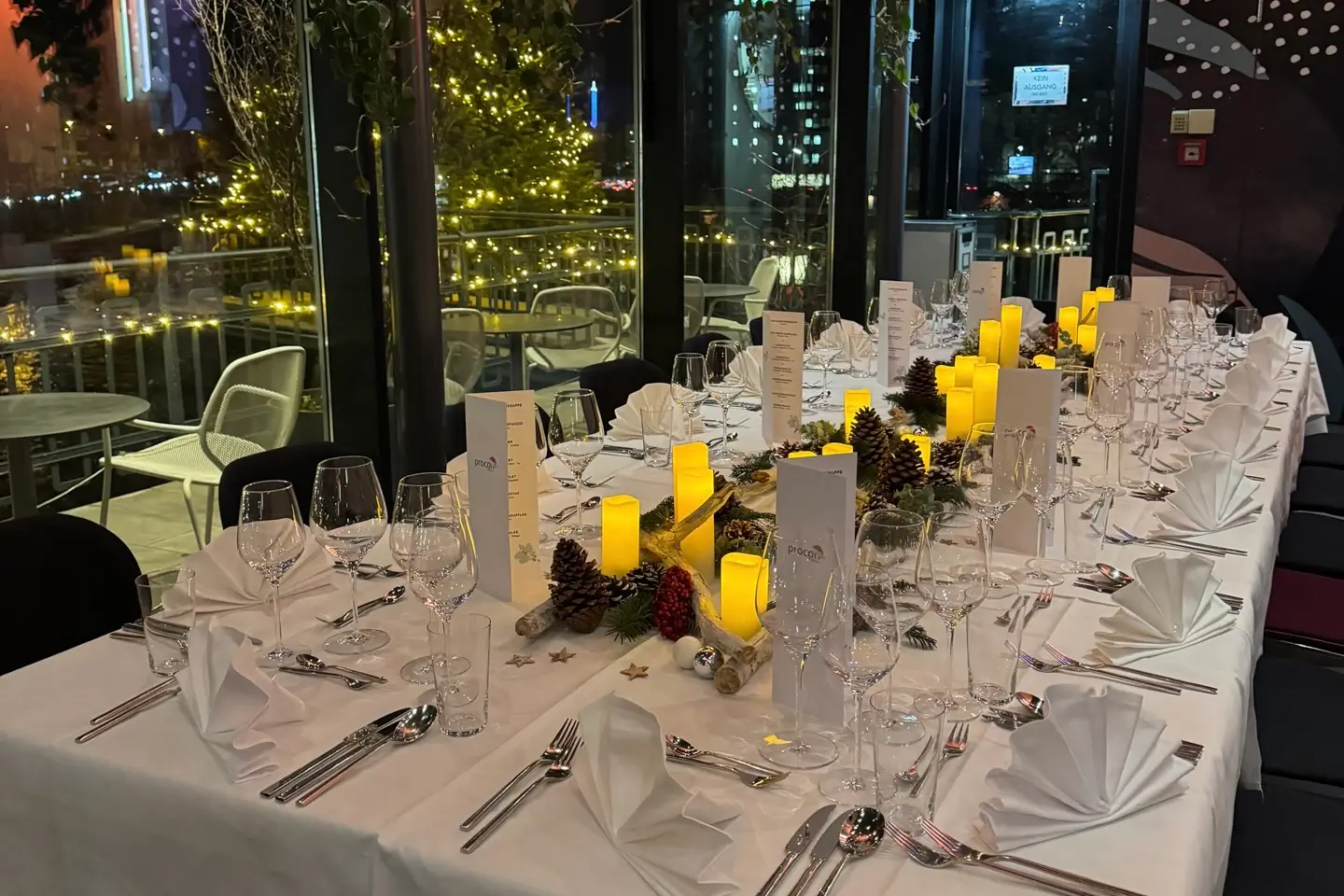 Ein langer Esstisch ist elegant mit weißer Bettwäsche, gefalteten Servietten, Gläsern, Silberbesteck, Tannenzapfen und leuchtenden Kerzen gedeckt, mit Blick auf die Lichter der Stadt und einen geschmückten Baum vor dem Fenster.