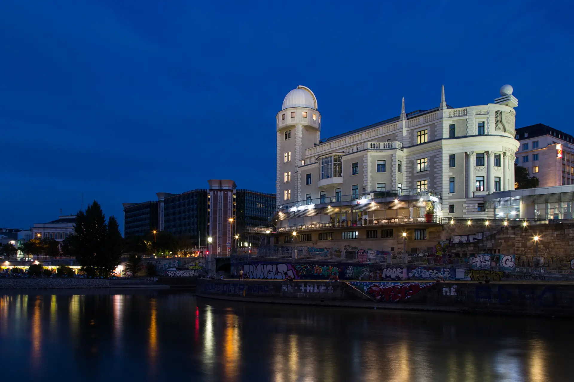 Ein großes, beleuchtetes Gebäude mit einer Kuppel steht nachts an einem Fluss und reflektiert das Licht auf dem Wasser. Die Uferwand ist mit bunten Graffiti beschmiert, und der Himmel ist tiefblau. Im Hintergrund sind moderne Gebäude zu sehen.