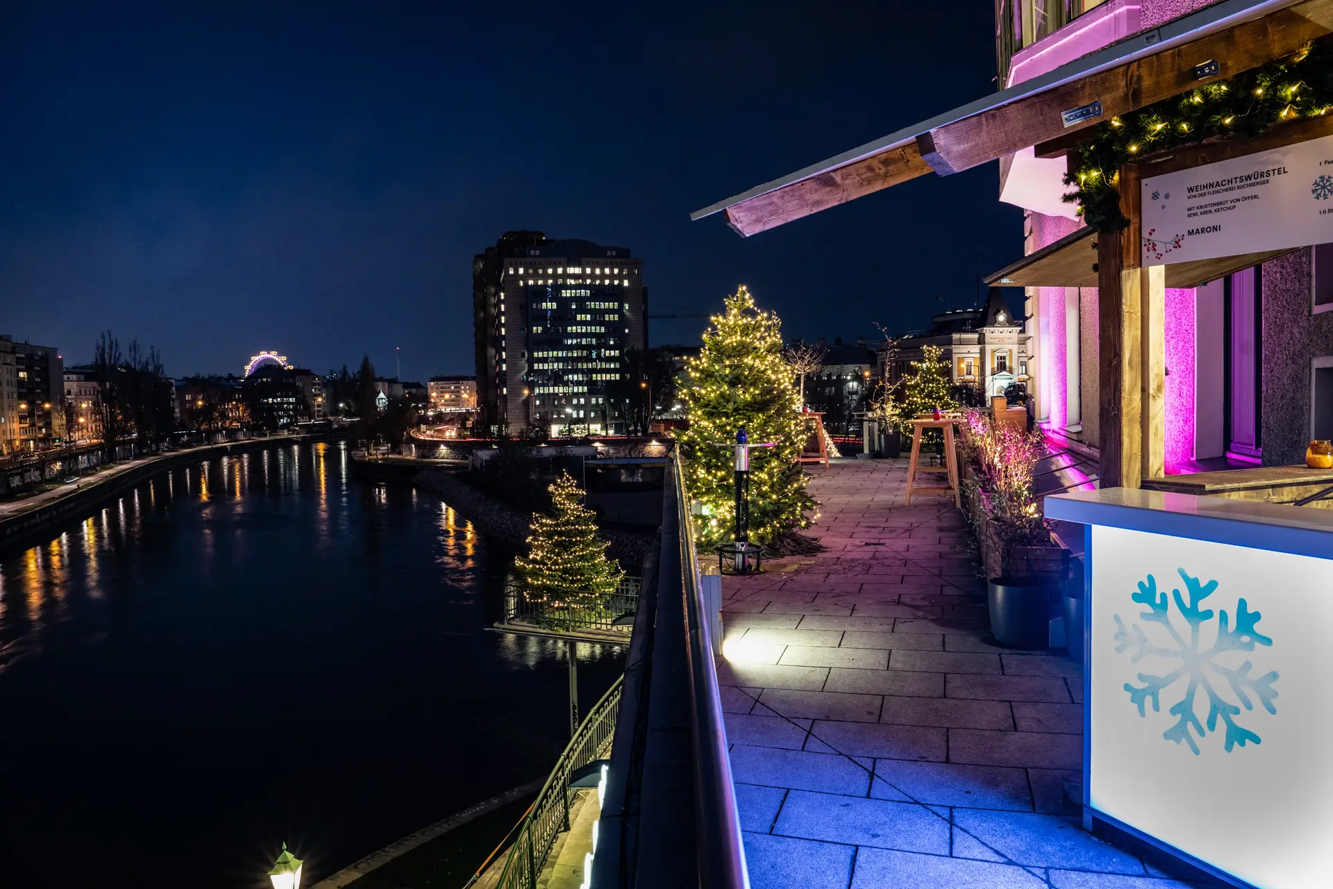 KLYO Eine festliche nächtliche Flussszene mit geschmückten Weihnachtsbäumen, beleuchteten Gebäuden und einer blau leuchtenden Theke mit Schneeflockenmuster, mit Blick auf das Wasser und die Lichter der Stadt im Hintergrund.