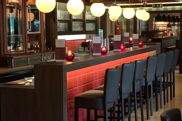 Eine moderne Bar mit hohen Stühlen, einer mit roten Kacheln ausgelegten Arbeitsplatte, darüber leuchtende runde Pendelleuchten und rote Kerzenhalter. Im Hintergrund sind Glasregale mit Gläsern und Flaschen zu sehen.
