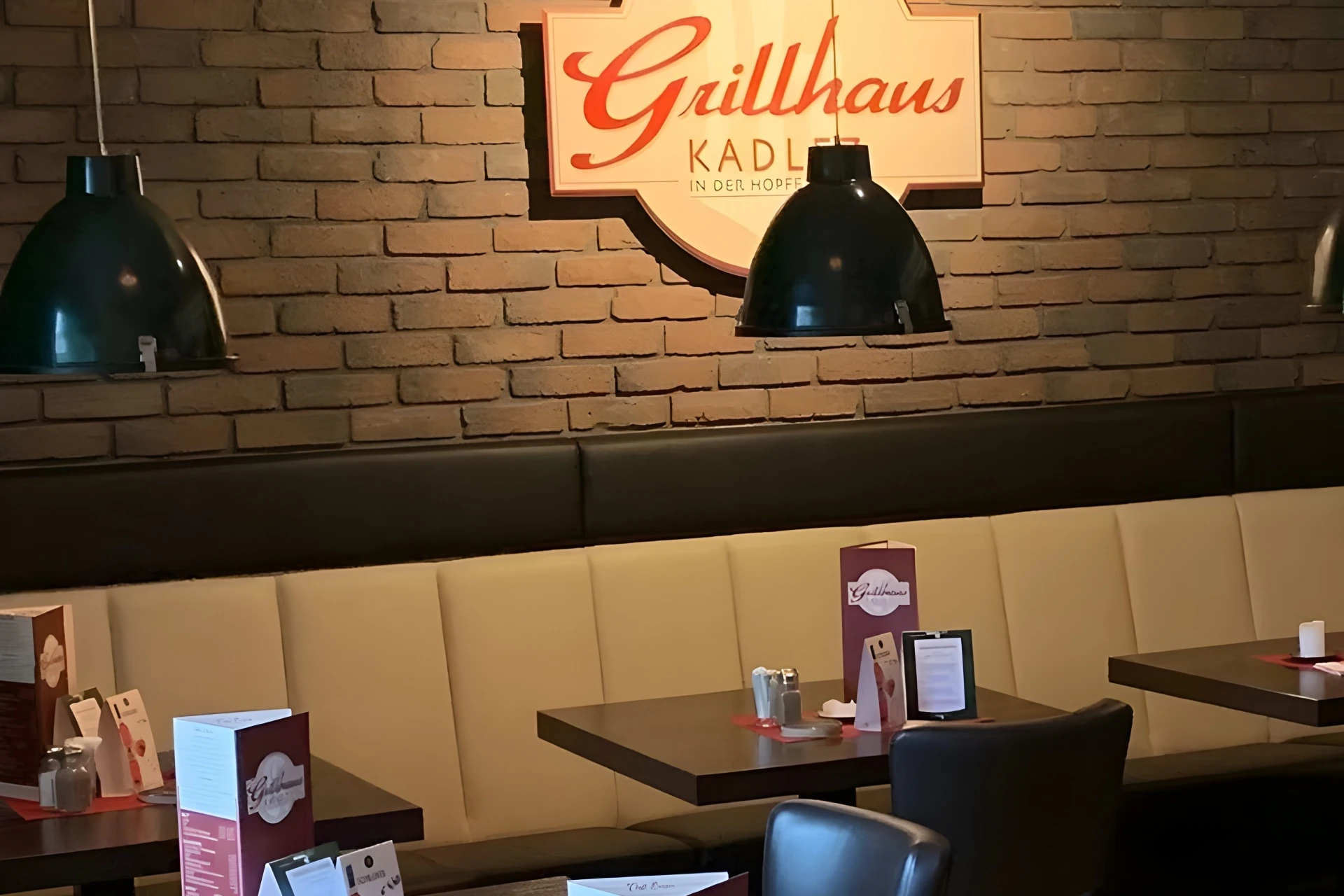 Ein gemütliches Restaurantinterieur mit cremefarbenen Sitzbänken, dunklen Holztischen und schwarzen Hängelampen. An der Backsteinwand hängt ein großes Schild mit der Aufschrift Grillhaus KADL. Speisekarten und Gewürze stehen auf den Tischen.