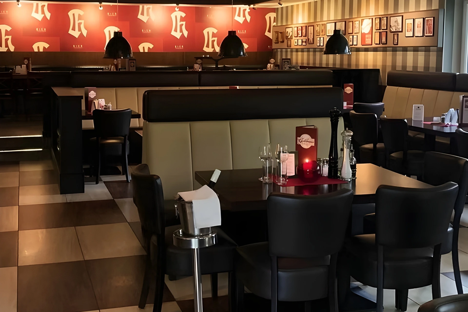 Ein modernes Restaurant mit schwarzen Stühlen, Holztischen mit Gläsern und einem rot-beigen Farbschema. An den Wänden hängen gerahmte Bilder und das Logo des Restaurants. Die sanfte Beleuchtung schafft eine gemütliche Atmosphäre.