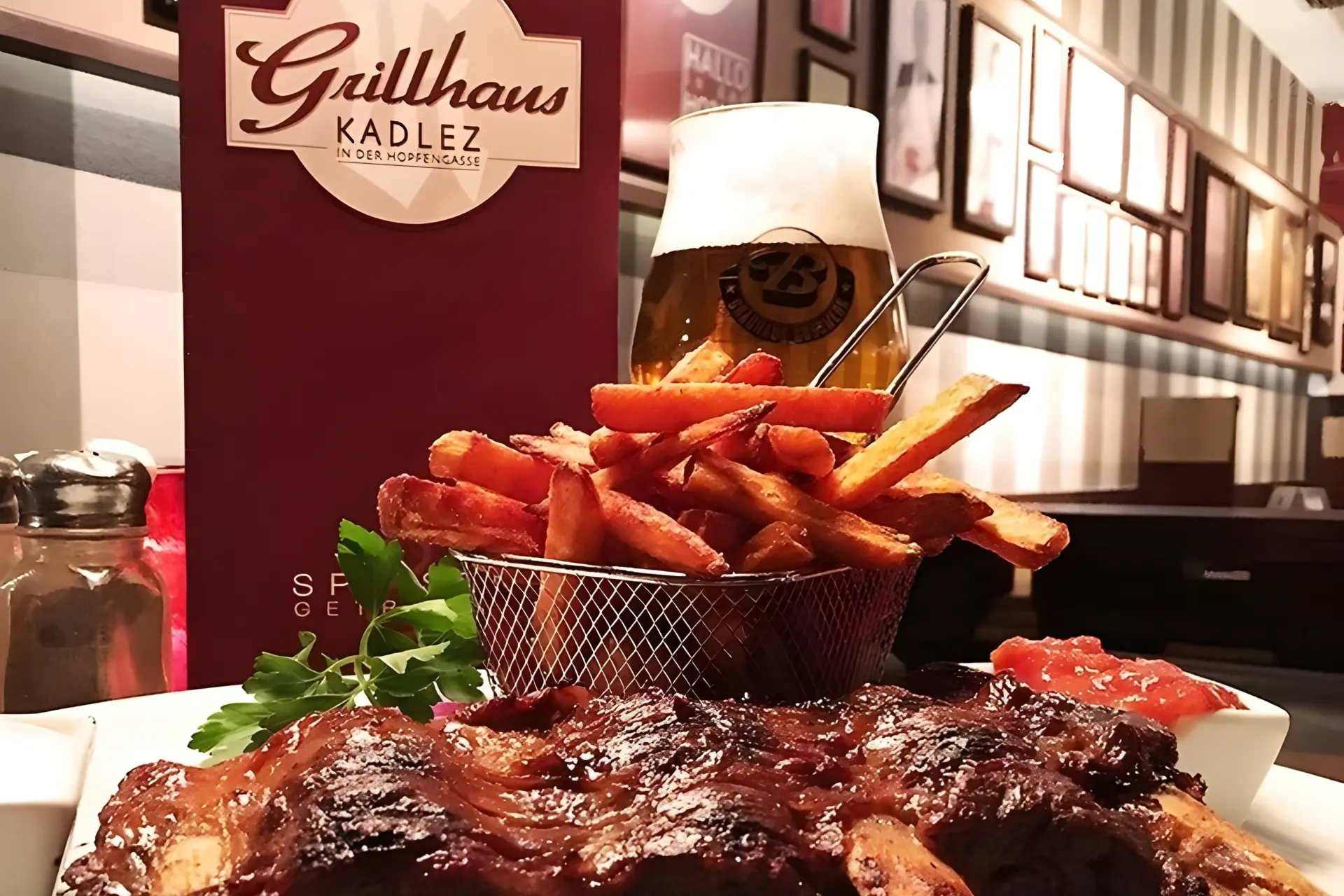 Ein Teller mit glasierten Rippchen, knusprigen Pommes frites in einem Metallkorb, einer Soße und einem Glas Bier, mit einer Grillhaus Kadlez Speisekarte und einem Salzstreuer im Hintergrund.