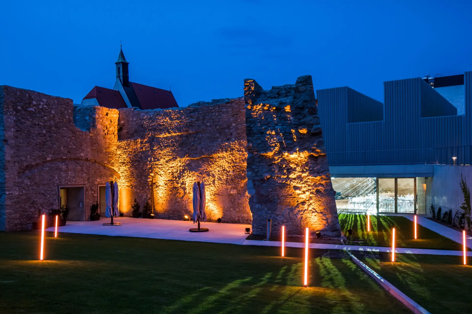 Die steinernen Ruinen und das moderne Kasematten-Gebäude werden in der Abenddämmerung von warmen Lichtern angestrahlt, während orangefarbene Pfähle eine Wiese säumen und im Hintergrund eine Kirche mit rotem Dach zu sehen ist.