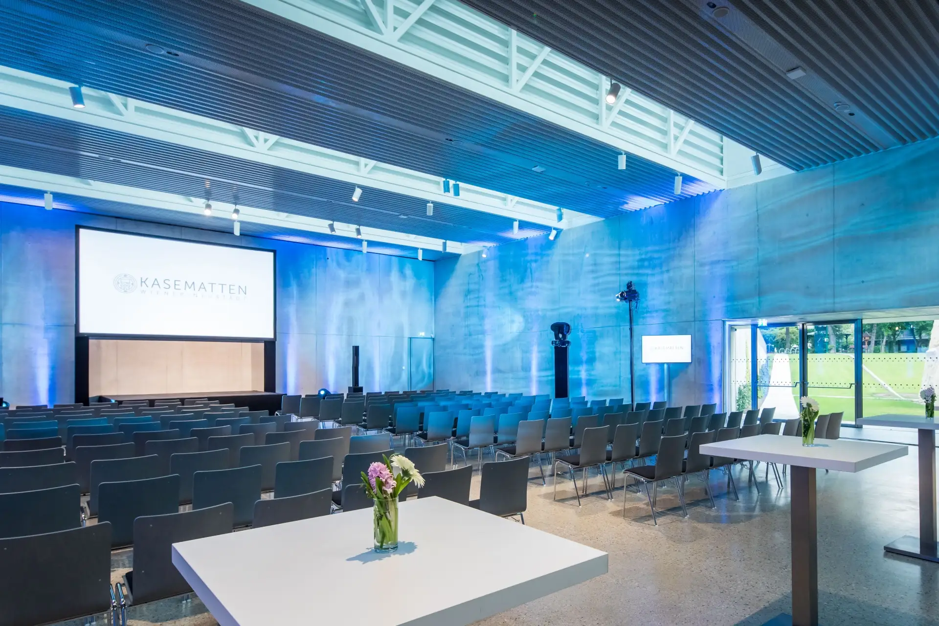 Moderner Konferenzraum in den Kasematten mit Stuhlreihen vor einer großen Projektionsfläche, blauer Akzentbeleuchtung an den Betonwänden, hohen Decken mit Sichtbalken und Tischen mit Blumenarrangements im Vordergrund.