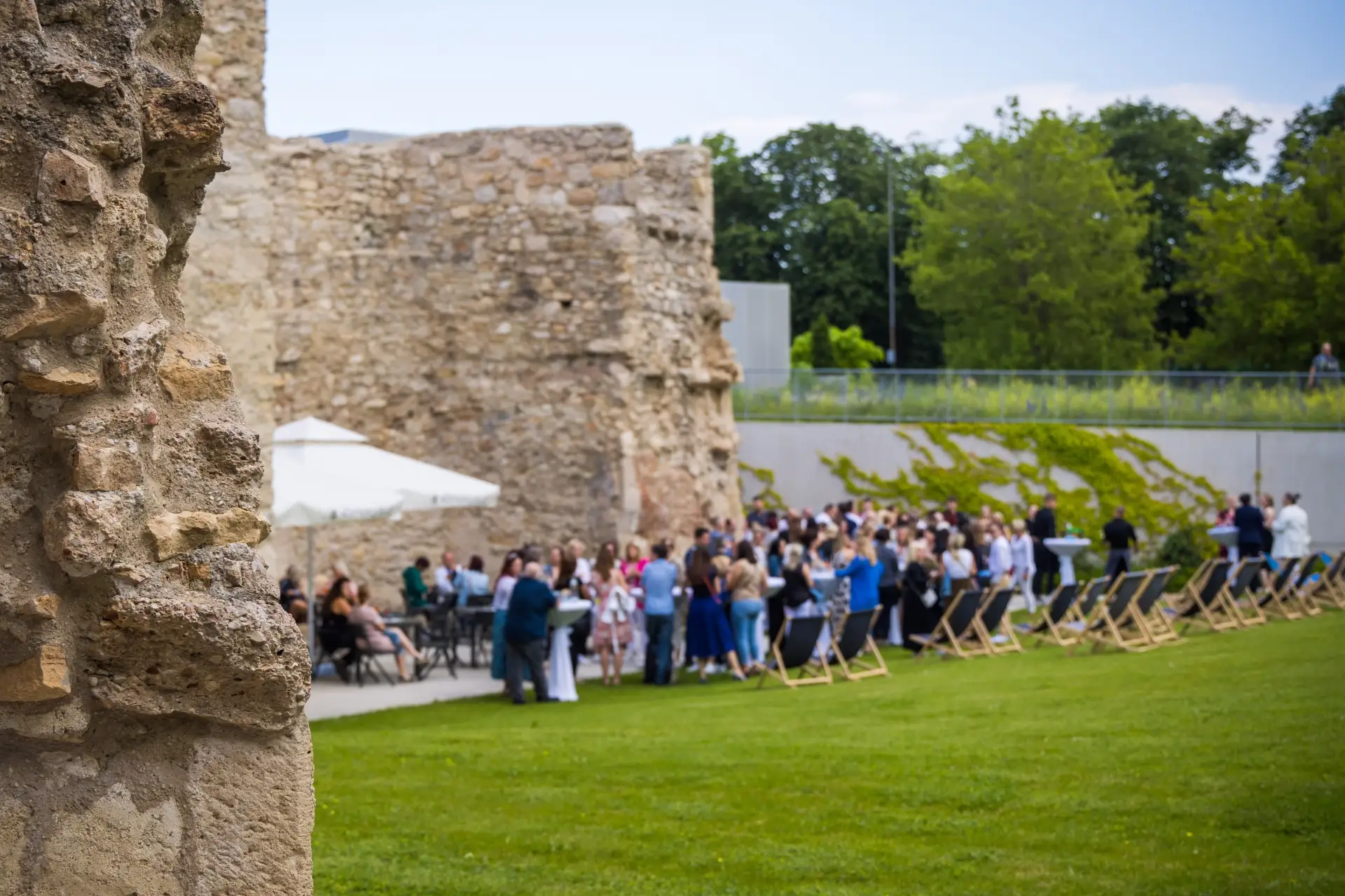 Eine Gruppe von Menschen versammelt sich im Freien in der Nähe der Steinruine Kasematten, wo Stühle und Tische im Gras aufgestellt sind. Im Hintergrund sind Bäume und eine Steinmauer zu sehen, und einige Menschen stehen unter einem weißen Sonnenschirm.