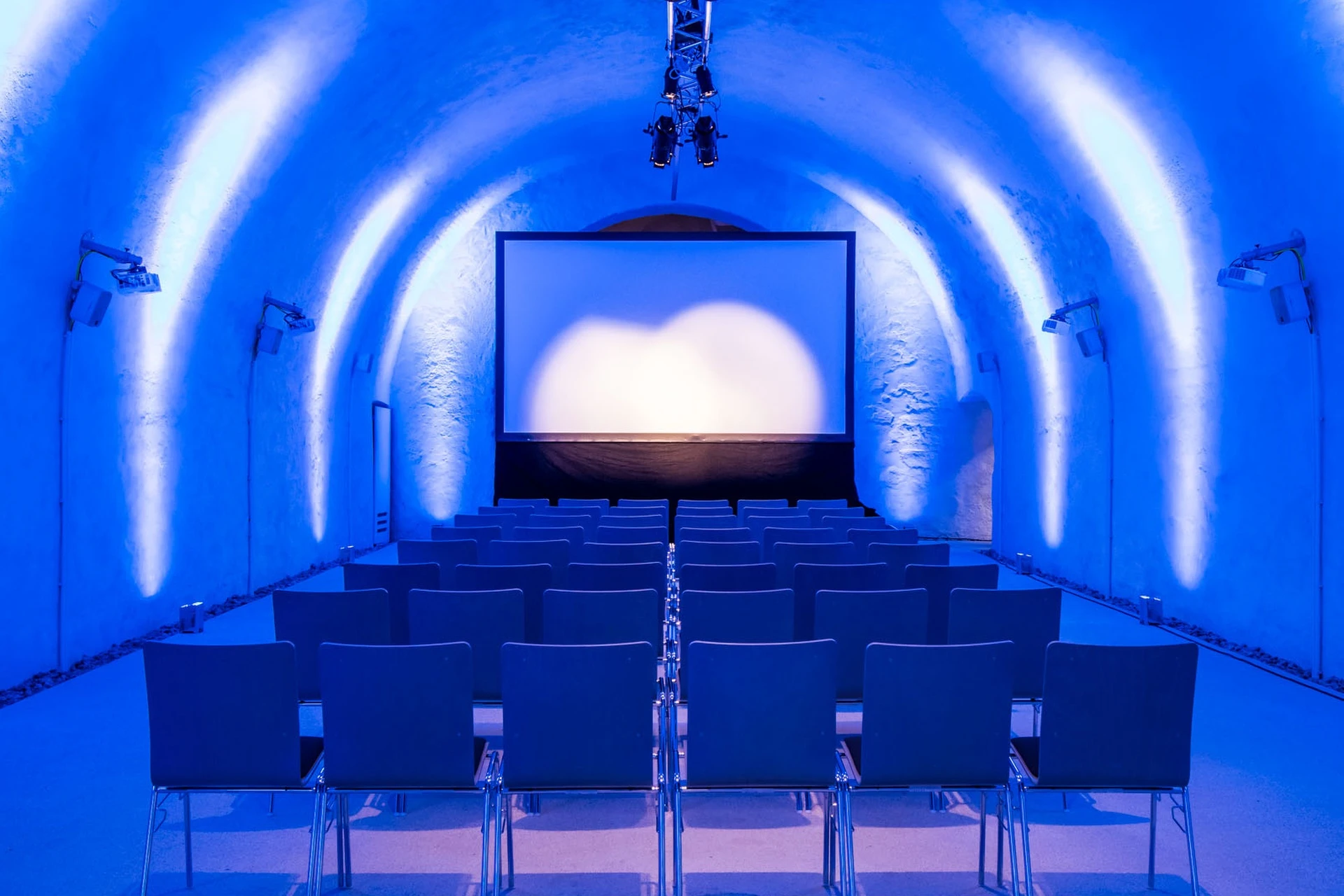 In den gewölbten, tunnelförmigen Kasematten stehen leere Stuhlreihen vor einer großen Leinwand, die mit blauem Licht angestrahlt wird und einen modernen und stimmungsvollen Rahmen für eine Präsentation oder Veranstaltung schafft.