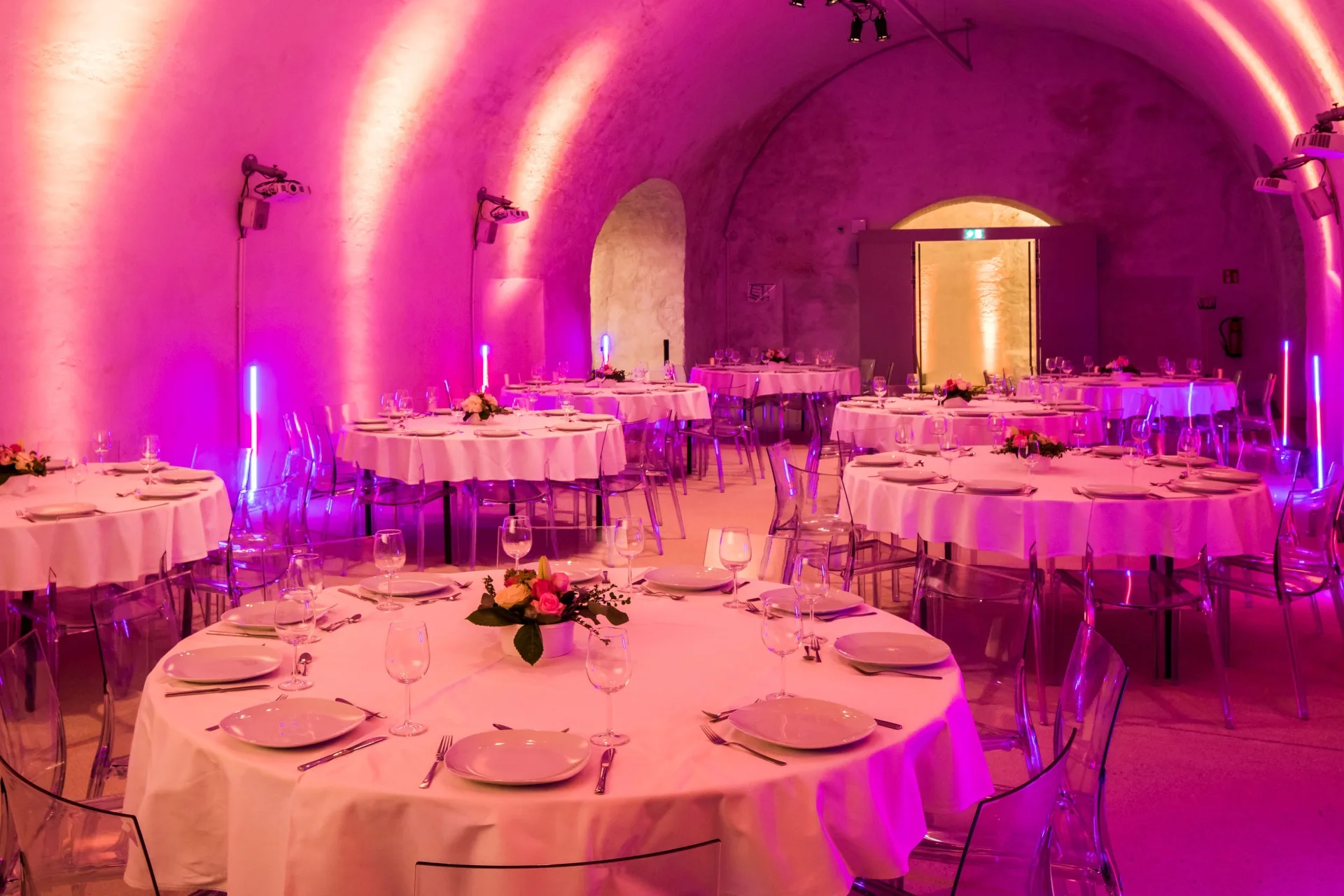 Runde Tische mit weißen Tischdecken, gedeckt mit Tellern, Gläsern und floralen Tafelaufsätzen, füllen den sanft beleuchteten Kasematten-Saal. Gewölbte Decken und rosa-violette Umgebungsbeleuchtung ergänzen die transparenten Stühle um jeden Tisch.