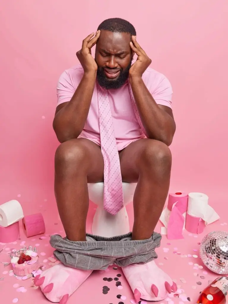 Ein Mann in rosa Hemd, Krawatte und Hausschuhen sitzt auf einer Toilette, den Kopf in den Händen, und leidet offensichtlich an einem Kater. Der rosafarbene Hintergrund ist mit Toilettenpapier, Konfetti und Partyutensilien übersät und fängt die Nachwirkungen einer wilden Nacht ein.