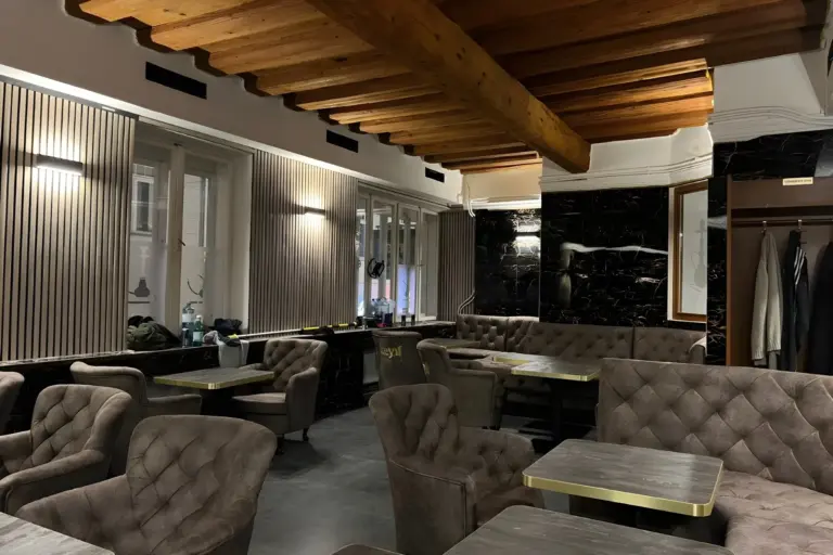 Ein modernes, leeres Café mit getufteten grauen Stühlen, Holztischen, einer Akzentwand aus dunklem Marmor und freiliegenden Holzbalken an der Decke. Natürliches Licht fällt durch große Fenster an einer Wand ein.