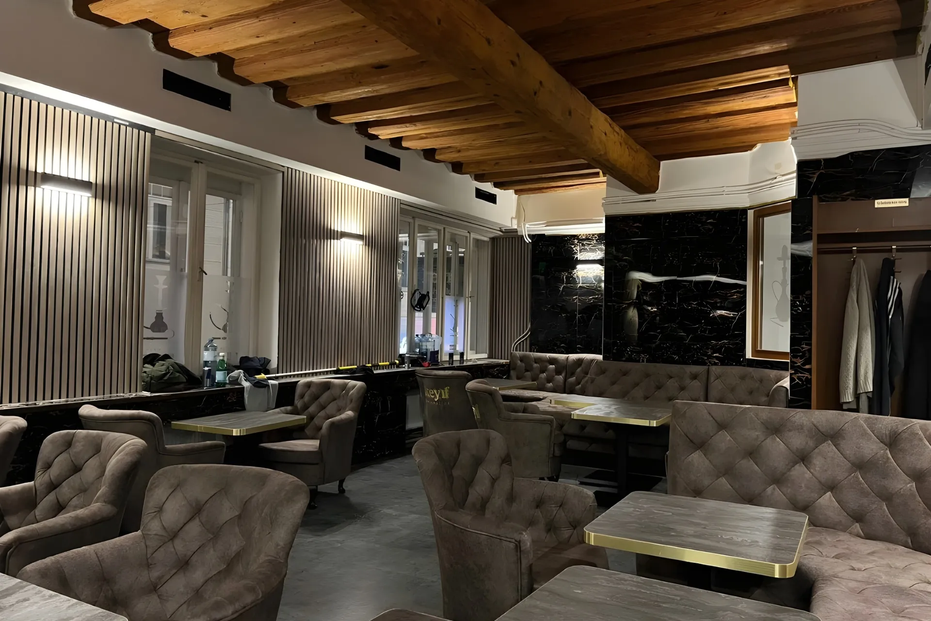 Ein modernes, leeres Café mit getufteten grauen Stühlen, Holztischen, einer Akzentwand aus dunklem Marmor und freiliegenden Holzbalken an der Decke. Natürliches Licht fällt durch große Fenster an einer Wand ein.