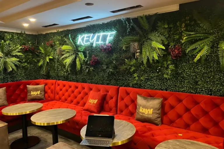 Eine gemütliche Lounge mit einem roten, getufteten Sofa, runden Marmortischen und einer üppig begrünten Wand mit Pflanzen und einer Leuchtreklame mit der Aufschrift KEYIF. Ein Laptop steht aufgeklappt auf einem Tisch, und auf dem Sofa liegen Markenkissen.