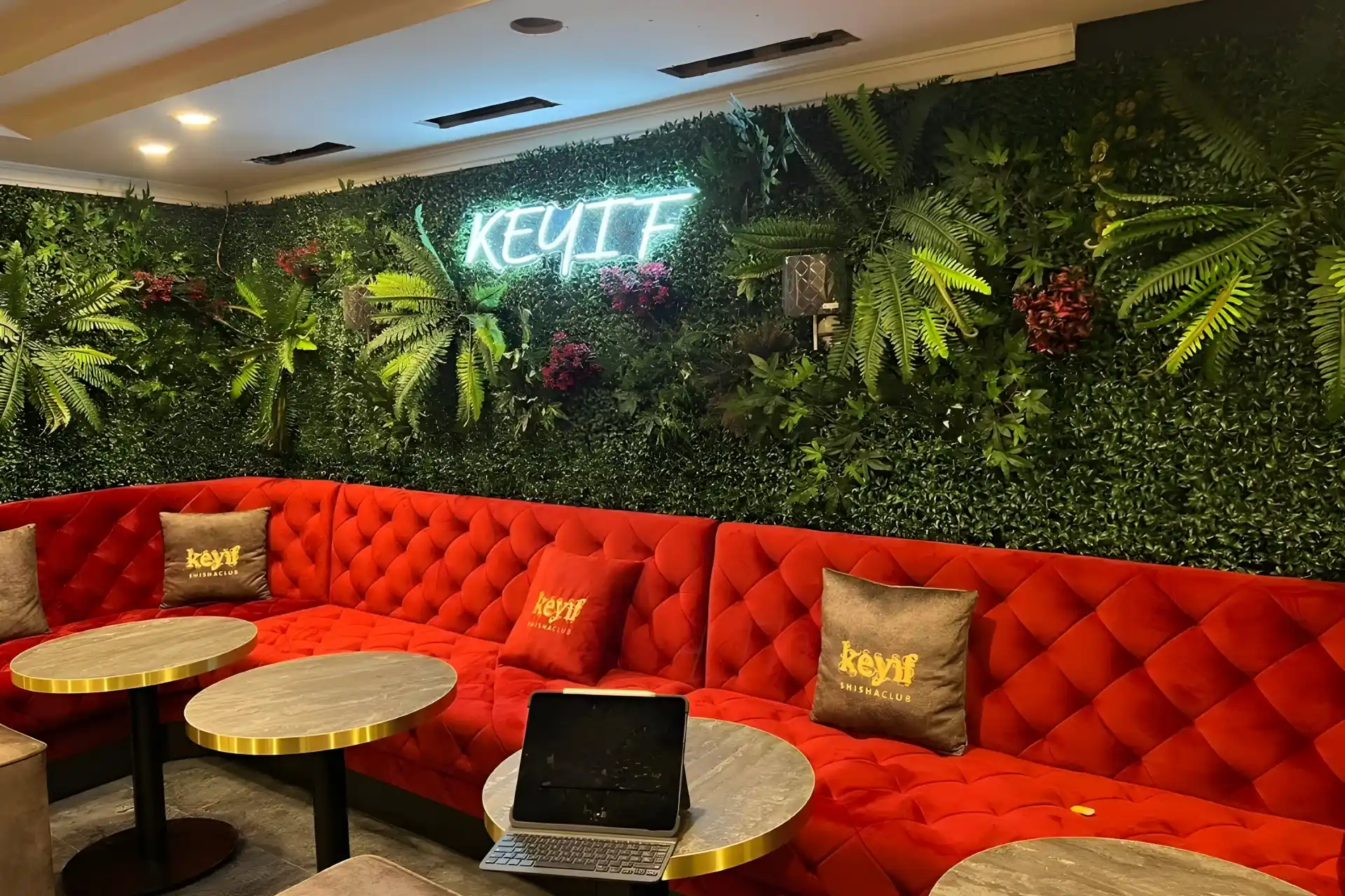 Eine gemütliche Lounge mit einem roten, getufteten Sofa, runden Marmortischen und einer üppig begrünten Wand mit Pflanzen und einer Leuchtreklame mit der Aufschrift KEYIF. Ein Laptop steht aufgeklappt auf einem Tisch, und auf dem Sofa liegen Markenkissen.