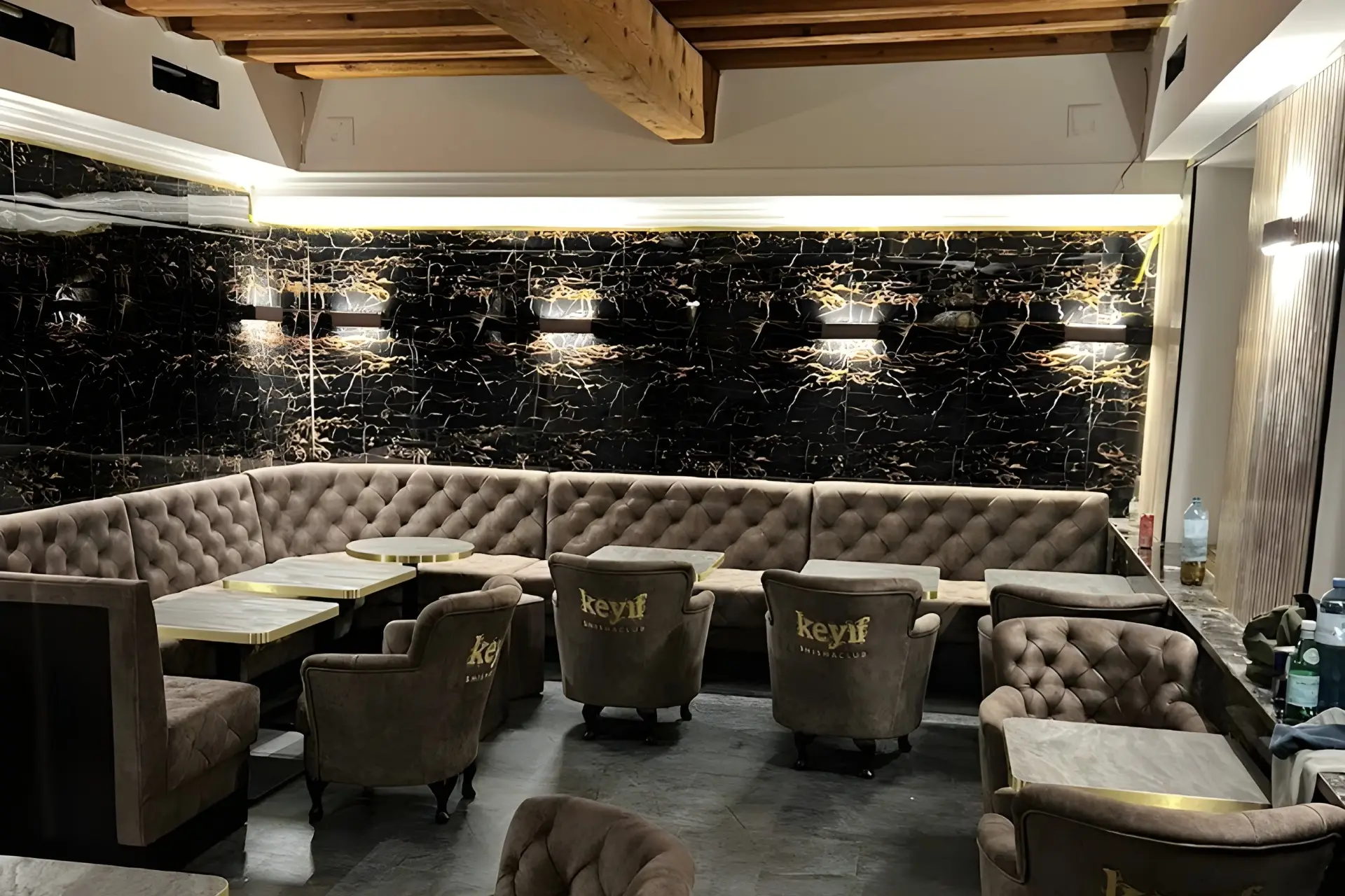 Eine moderne Lounge mit getufteten braunen Stühlen und Sofas, die um Holztische herum angeordnet sind, mit schwarzen Marmorwänden mit goldenen Akzenten und Deckenbalken. Sanfte Wandleuchten schaffen eine gemütliche Atmosphäre.