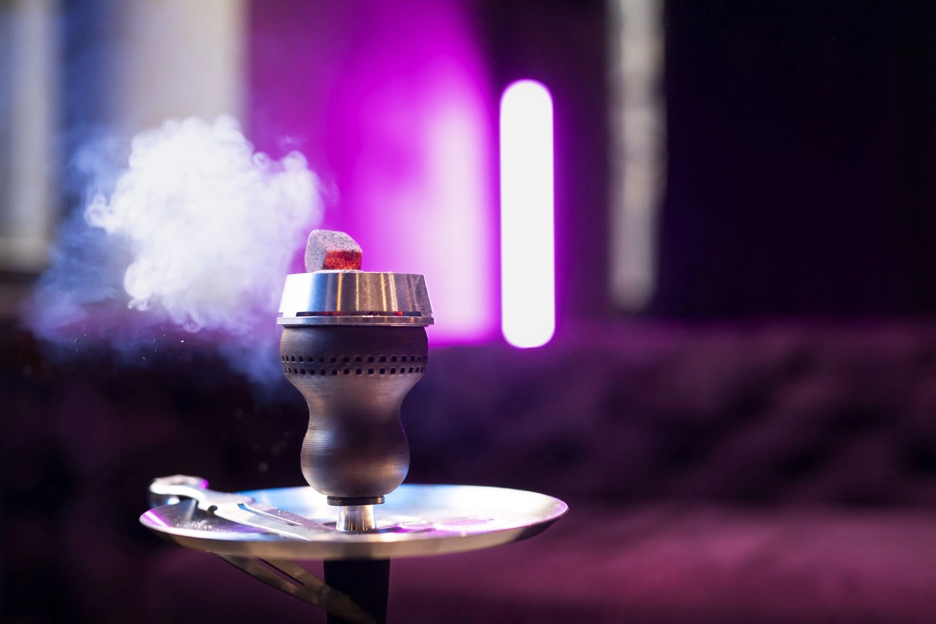 Eine angezündete Wasserpfeifenschale mit heißer Holzkohle sitzt auf einem Metalltablett und gibt Rauch ab. Der Hintergrund ist mit violetten und rosafarbenen Lichtern verschwommen und schafft eine entspannte und stimmungsvolle Atmosphäre.