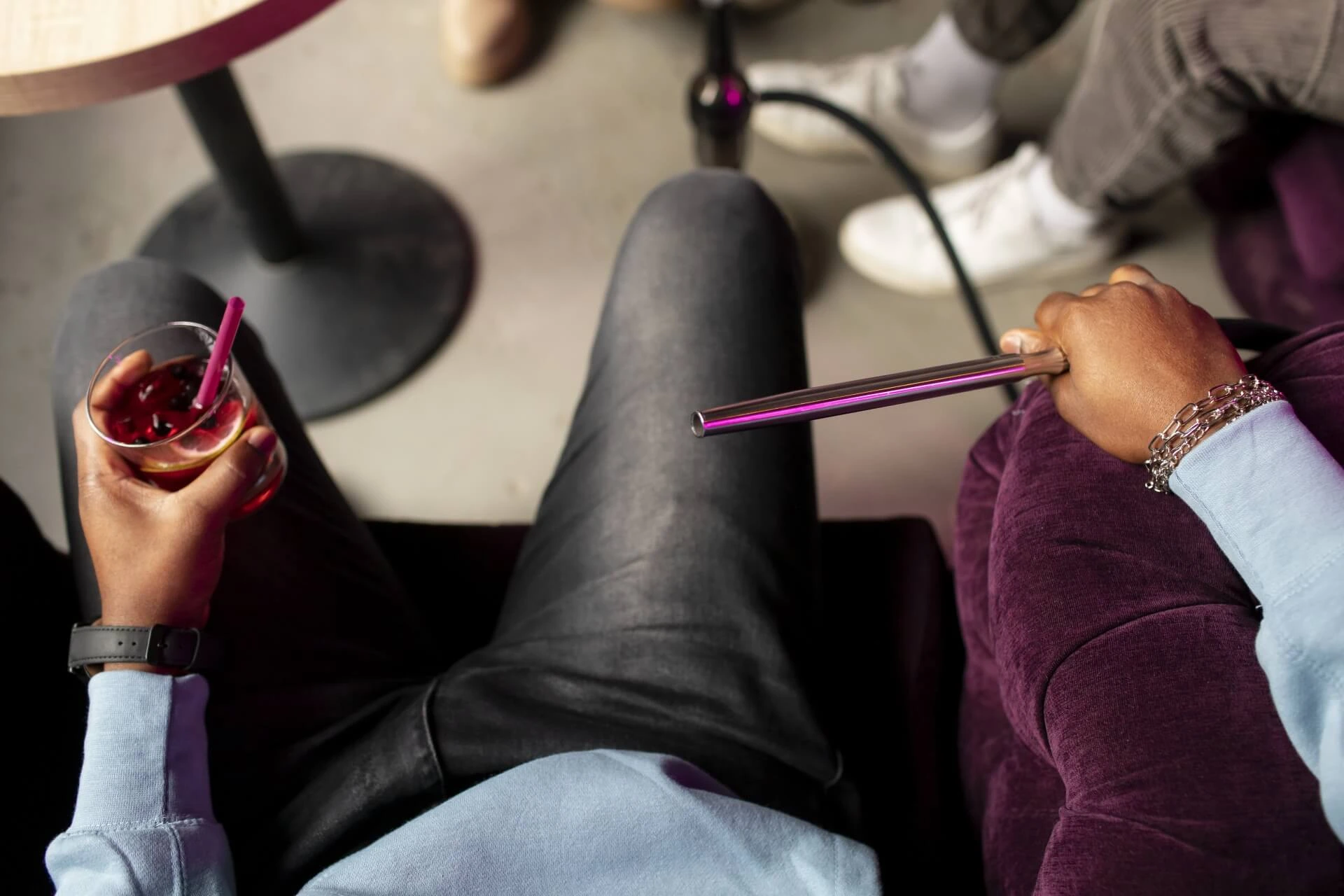 Auf einer lila Couch sitzende Person, die ein Getränk mit einem Strohhalm in der einen und einen Wasserpfeifenschlauch in der anderen Hand hält, wobei die Beine einer anderen Person und ein runder Tisch in der Nähe teilweise sichtbar sind.
